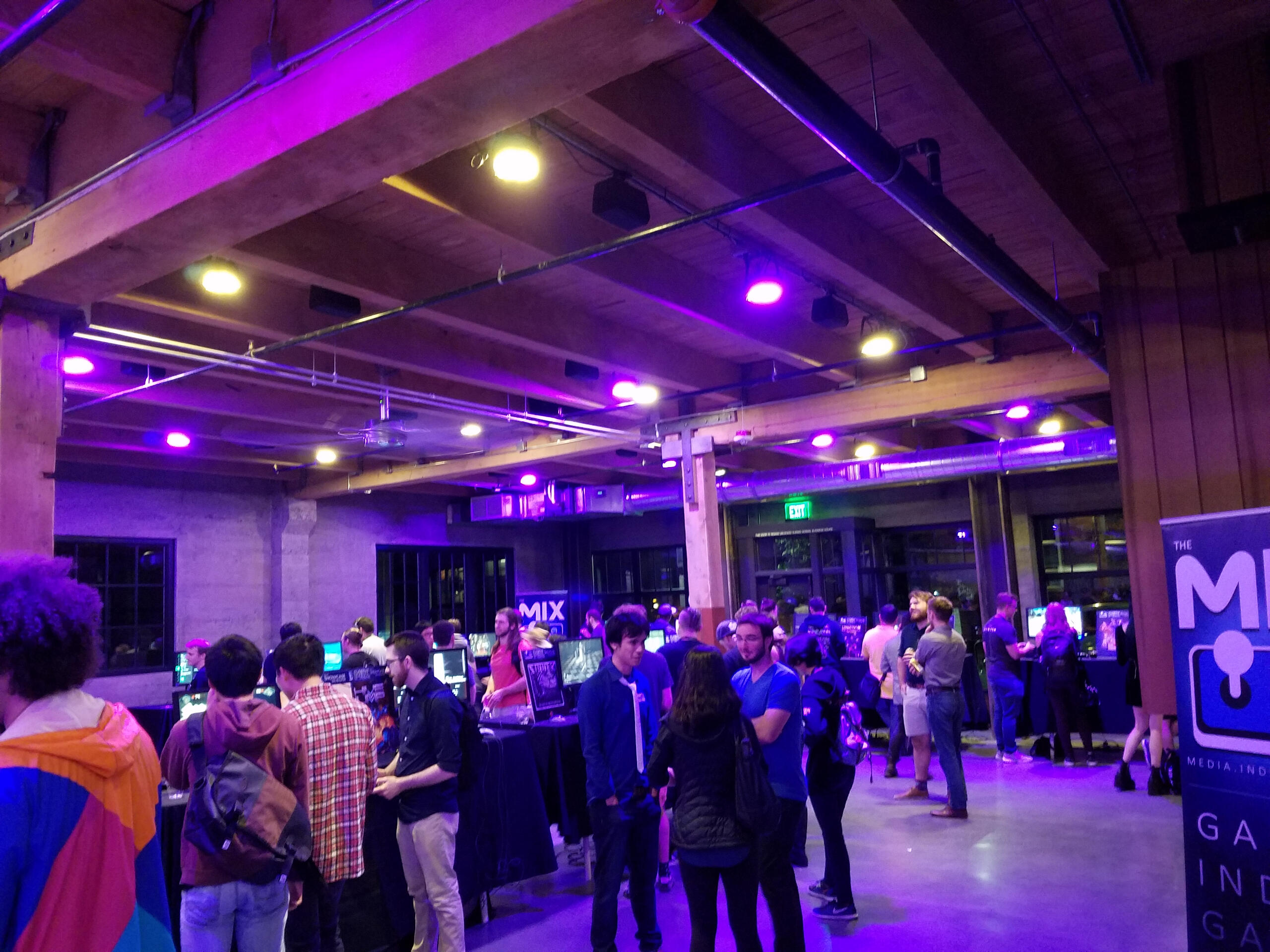 Past Event Image The picture shows an event bathed in purple light, where dozens of game devs and press stand around computers talking and playing demos of games.