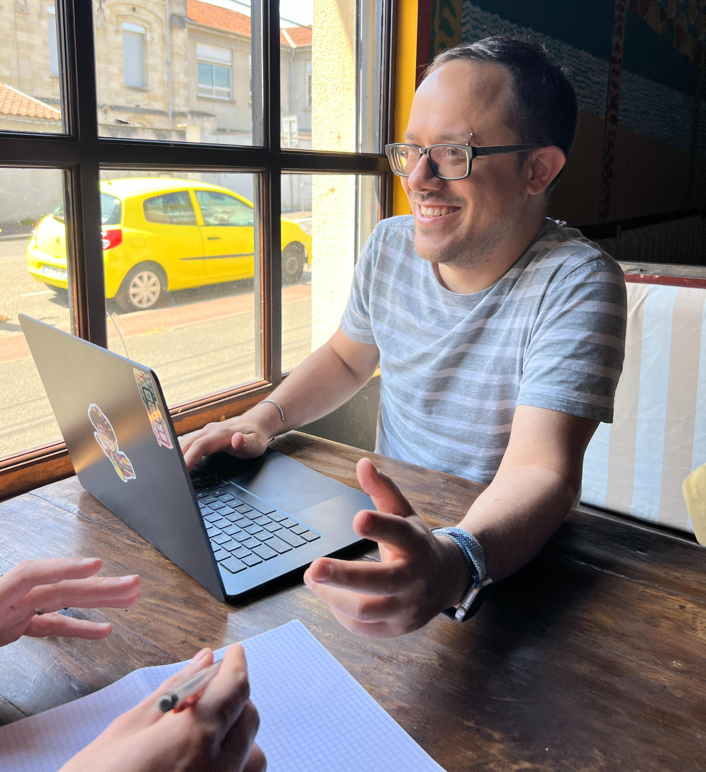 Coaching & Consulting Example Image of a white guy sitting at a booth in a restaurant using his computer. He's gesturing to someone mostly off camera. The hands of another person can be seen as well.